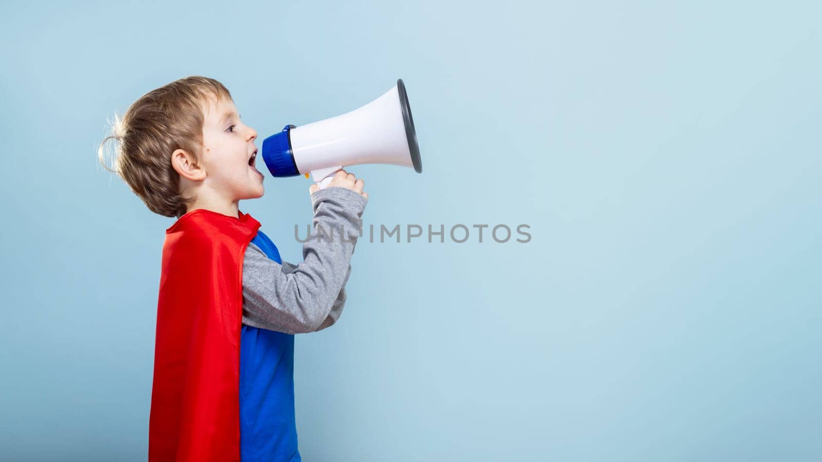 Young boy in superhero costume shouting into megaphone. Studio portrait with solid blue background. Empowerment and imagination concept. Design for banner, poster, invitation.