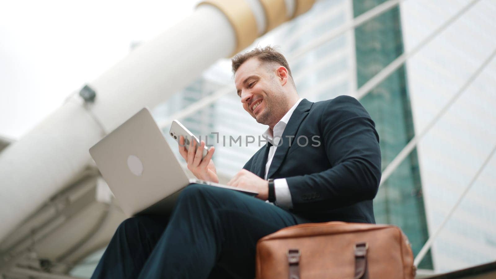 Caucasian business man sitting at stairs while using laptop and phone. Urbane. by biancoblue