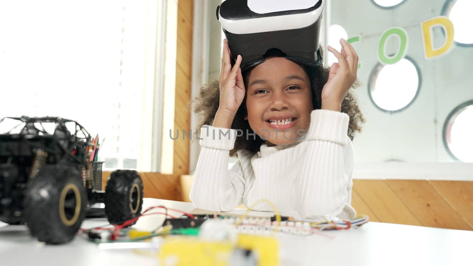 African girl taking off VR glass or head set while looking at camera. Student smiling at camera with colored pencil and laptop placed on table in STEM technology class. Online education. Erudition.