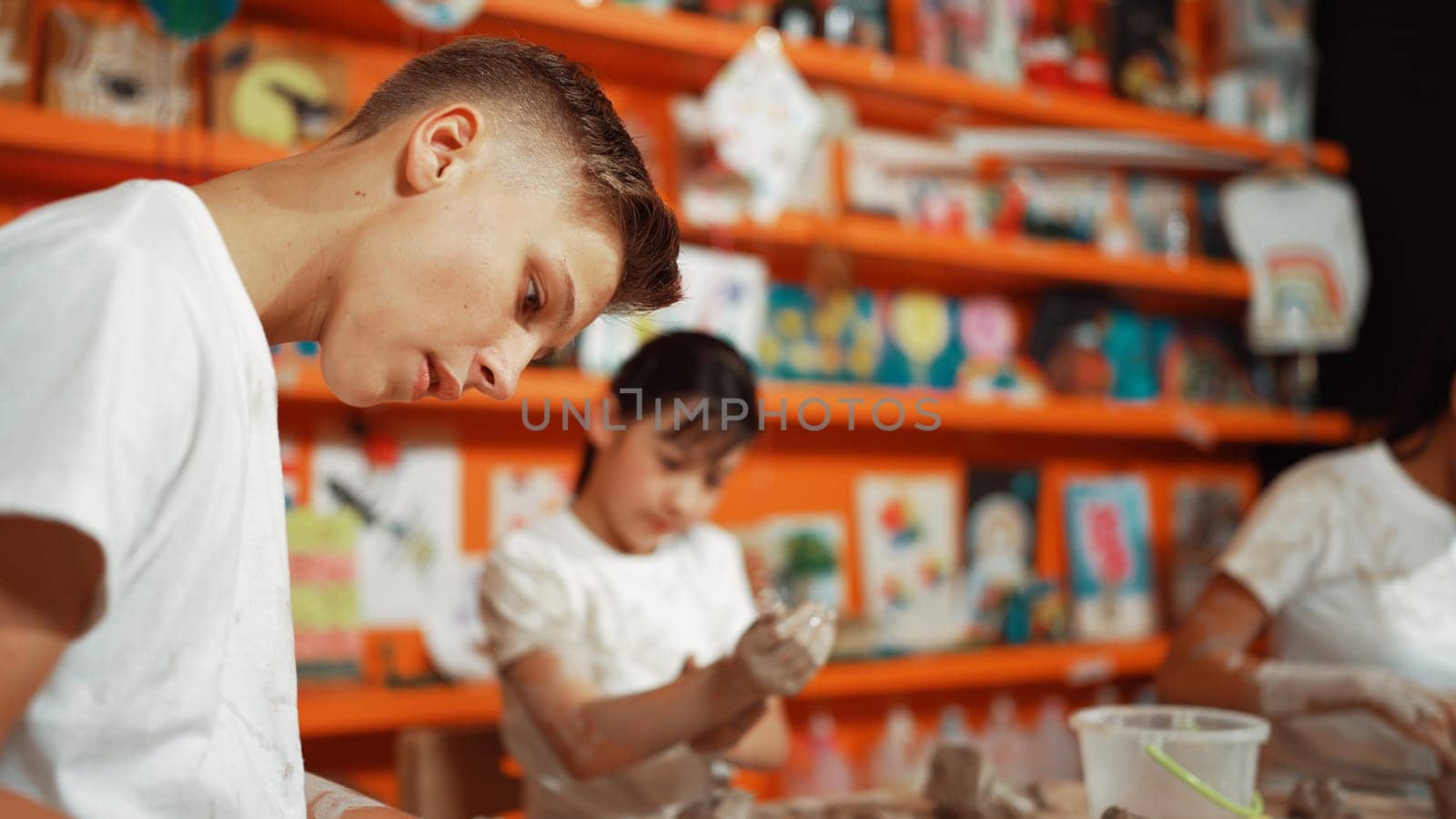 Asian highschool student modeling clay on table with mud and bottle of water at pottery workshop. Group of happy diverse children working or making cup of clay. Creative activity concept. Edification.