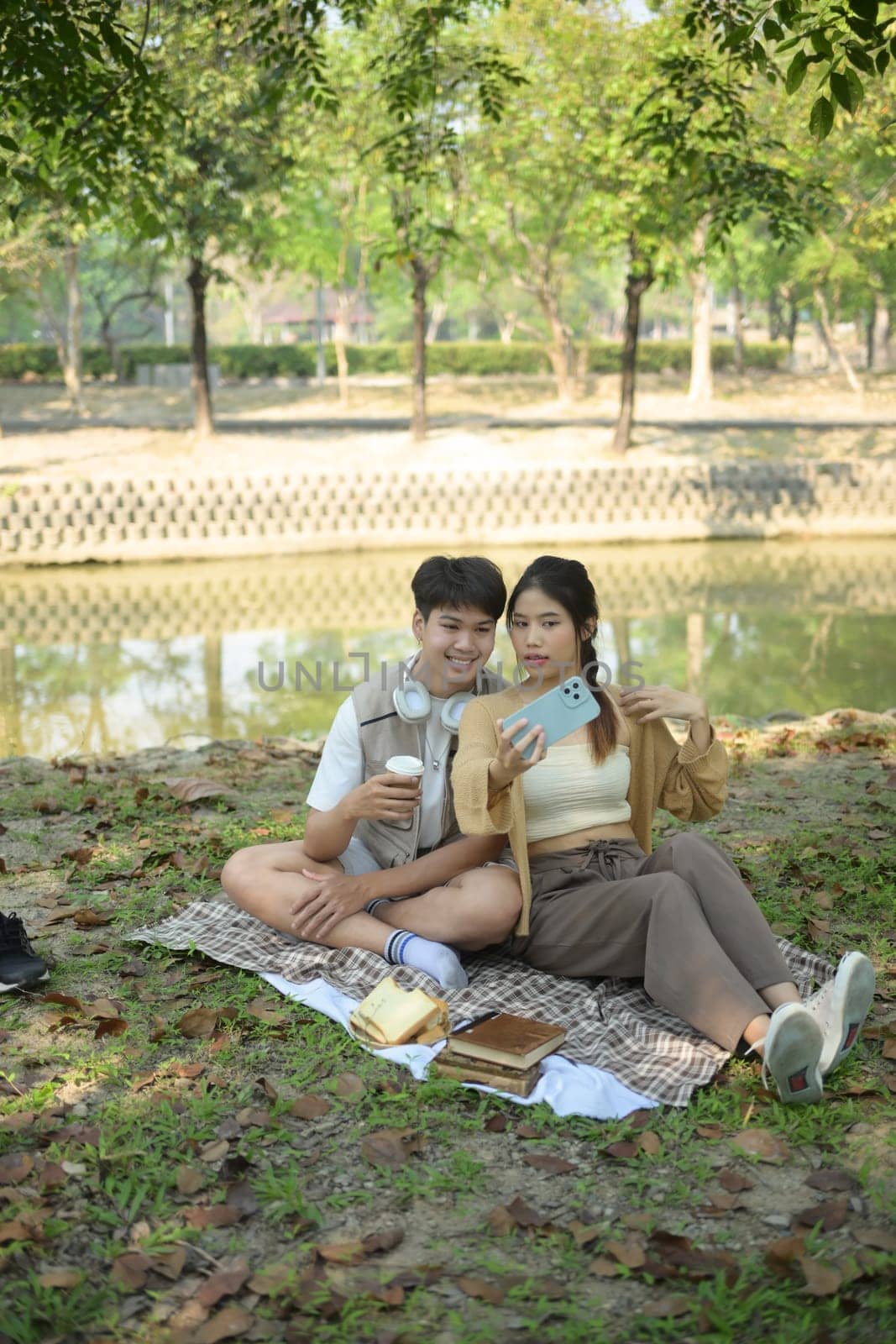 Couple in love taking selfie with smartphone, sitting on a picnic blanket in a public park by prathanchorruangsak