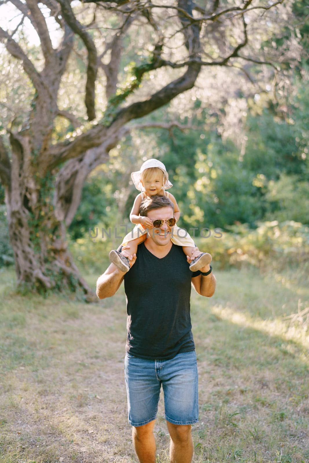 Little girl tugs sunglasses on dad face while sitting on his shoulders. High quality photo