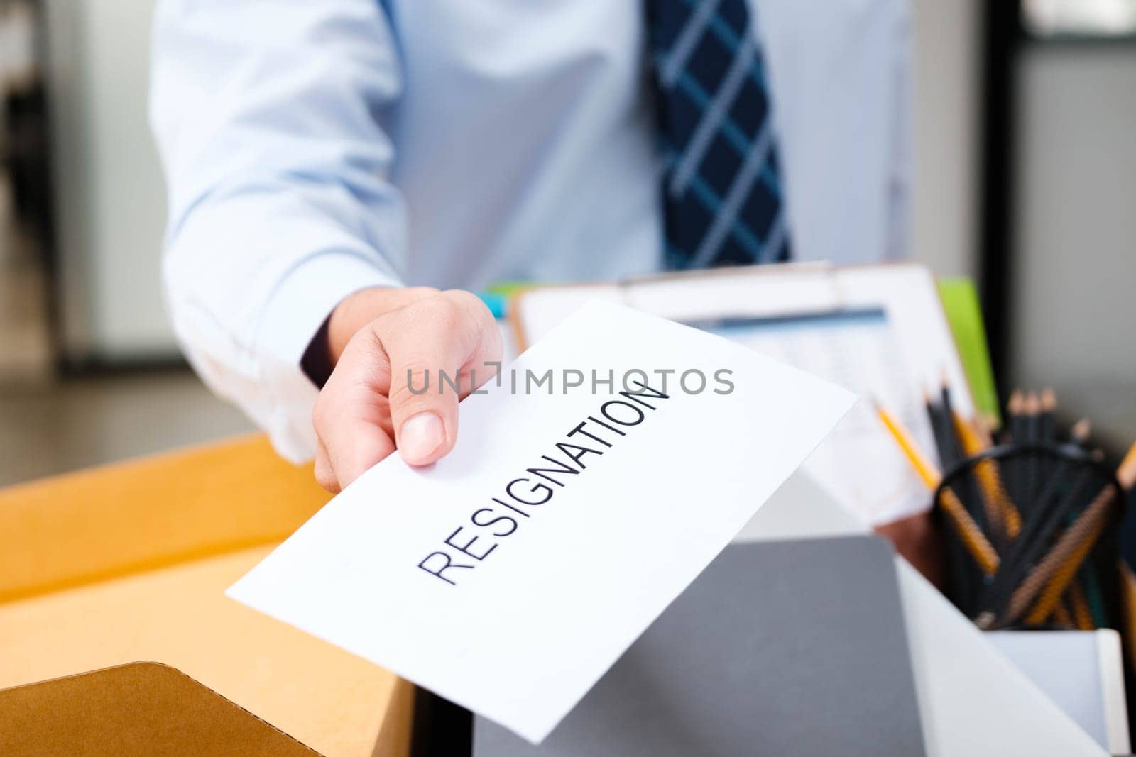 Close-Up of Man's Hand Delivering Resignation Letter with Box of Personal Belongings in the Background, Indicating Professional Departure