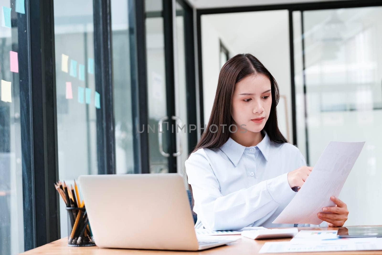 A focused female professional intently reviewing a document at her office desk with a laptop and smartphone nearby.