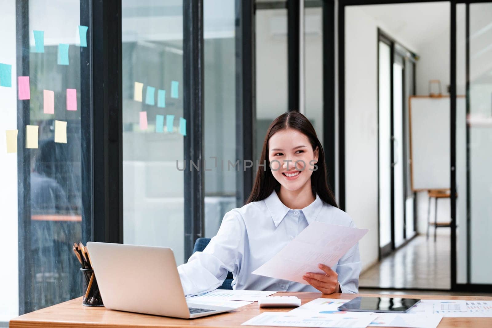 A cheerful female business professional reviewing documents with a laptop at her workspace in a well-lit office.