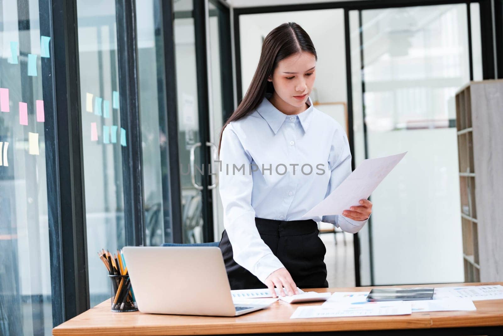 A focused female professional intently reviewing a document at her office desk with a laptop and smartphone nearby.