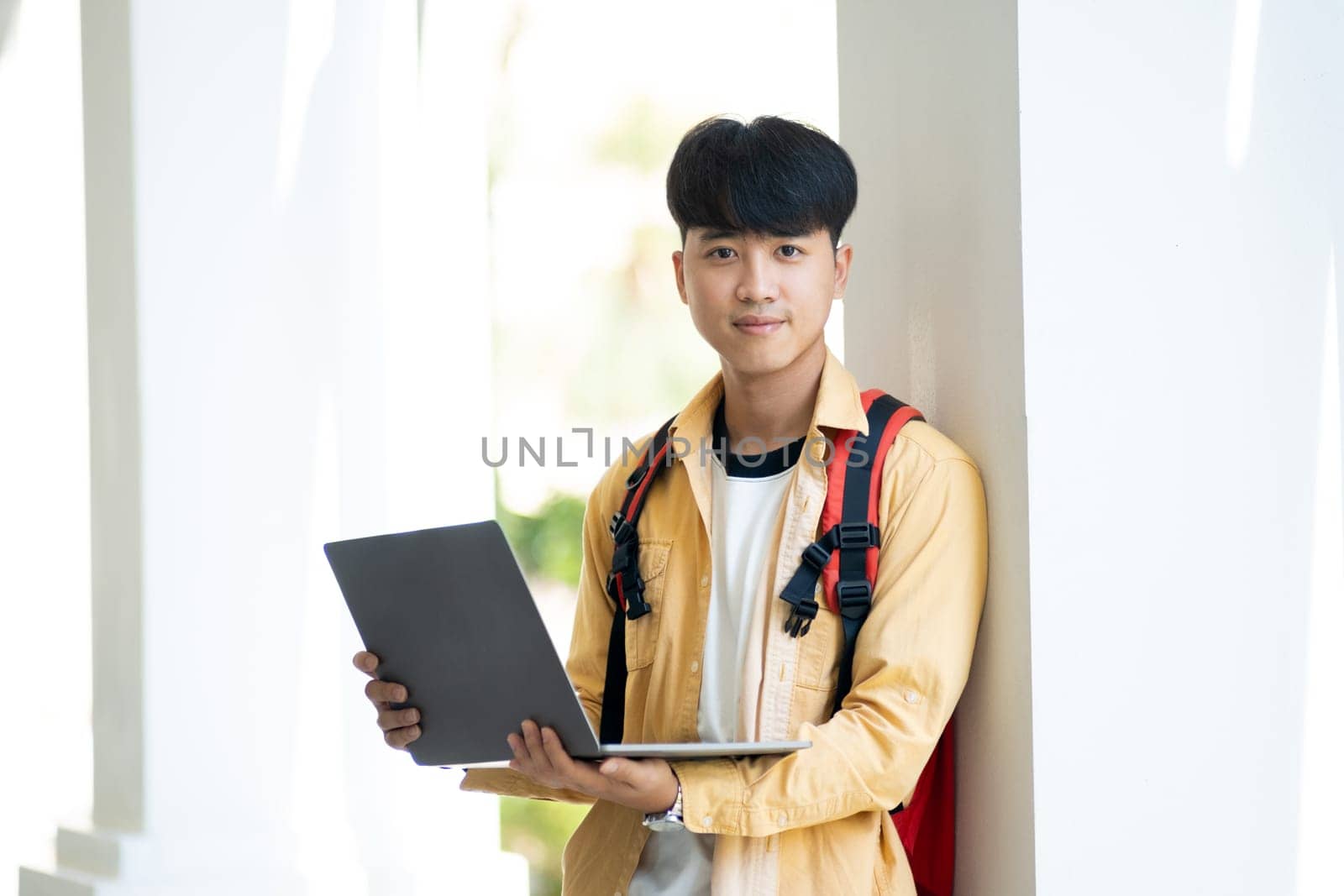 A male student with a pleasant smile stands in the school hallway, confidently holding his laptop, ready for class.