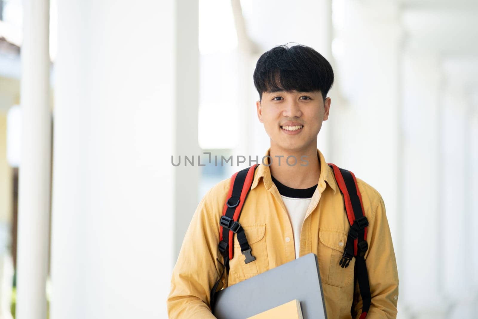 A college student stands in the hallway of his school, holding textbooks and smiling, ready for a day of learning and studies.