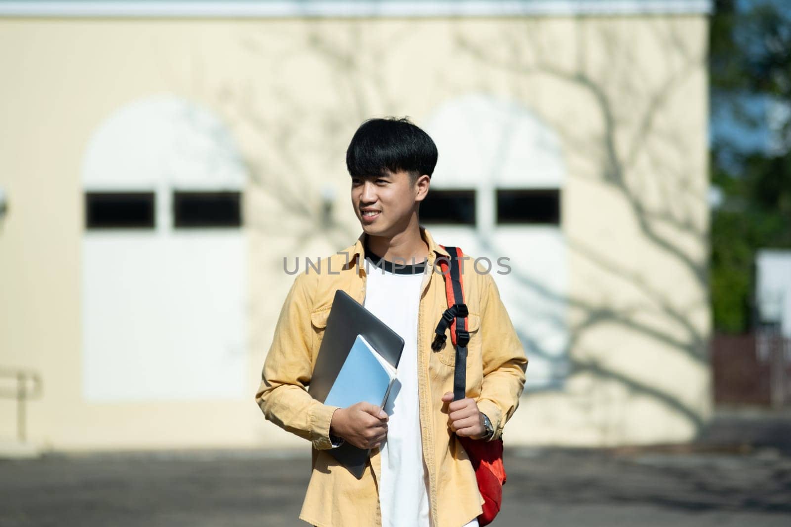A contented college student carrying a laptop and books walks across the campus grounds, exuding a sense of readiness and enthusiasm for learning.