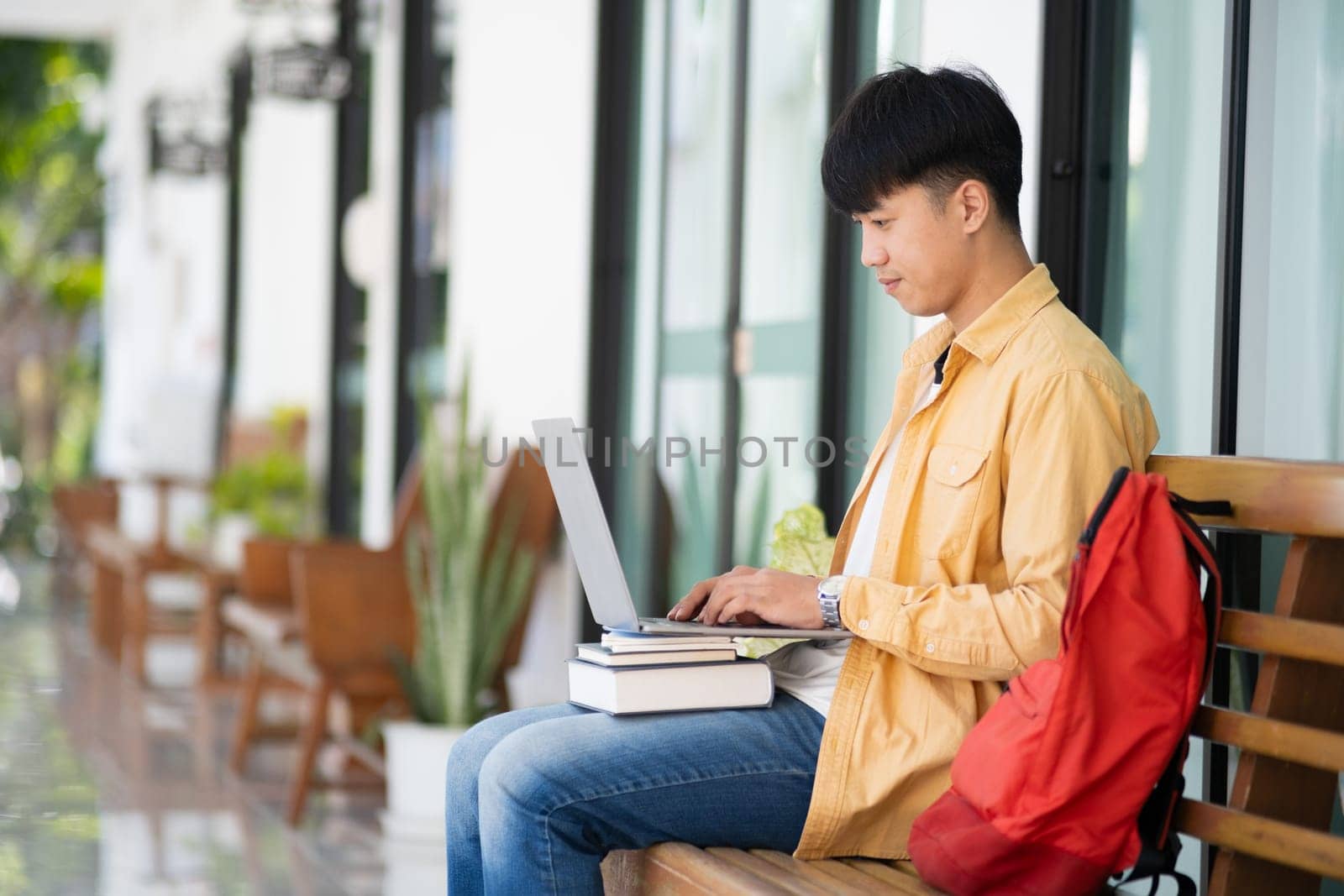 A college student sits on an outdoor bench, working intently on his laptop, with a stack of books and a red backpack by his side.