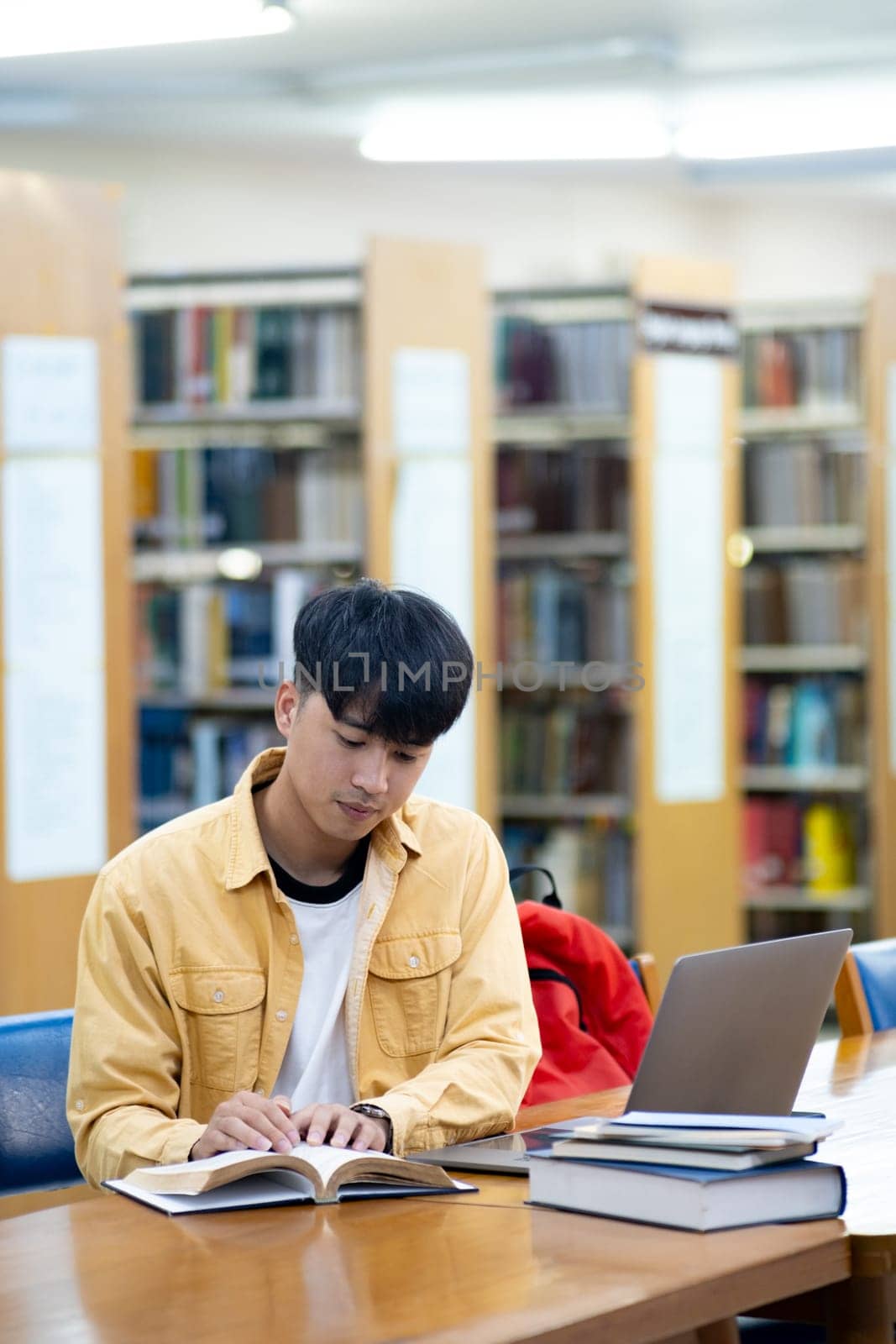 A young man is sitting at a table in a library, reading a book and using a laptop. Concept of focus and concentration, as the man is engaged in his studies. The library setting suggests a quiet