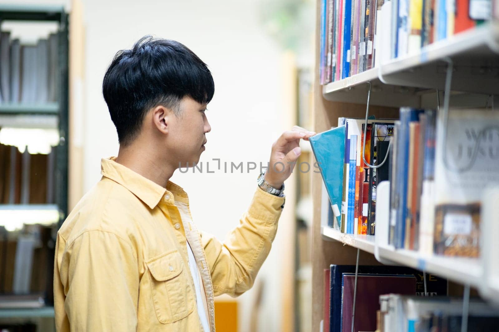 A man in a yellow shirt is looking at a book on a library shelf by ijeab
