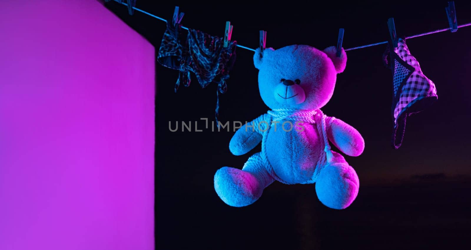 toy teddy bear in a bdsm mask and leather straps hangs on a clothesline in neon light on a dark background