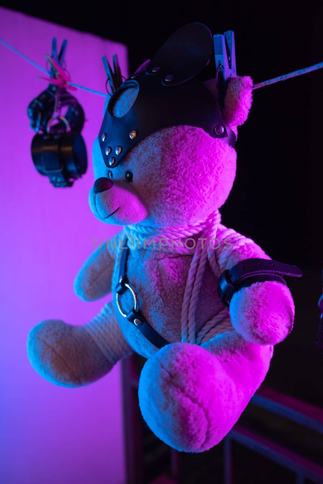 A toy teddy bear in a bdsm mask and leather straps hangs on a clothesline in neon light on a dark background by Rotozey