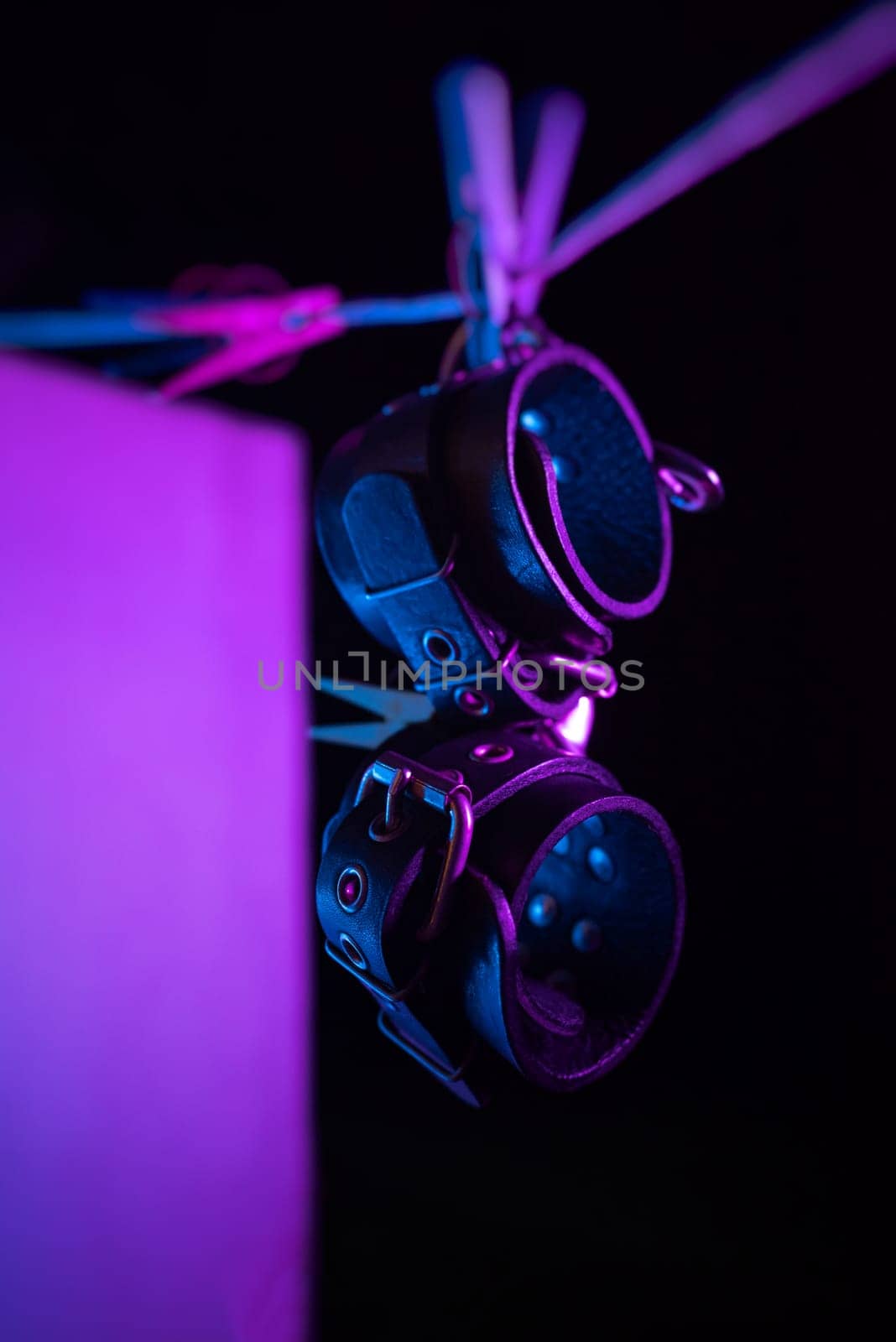 bdsm leather handcuffs hanging on a clothesline in neon light on a black background by Rotozey