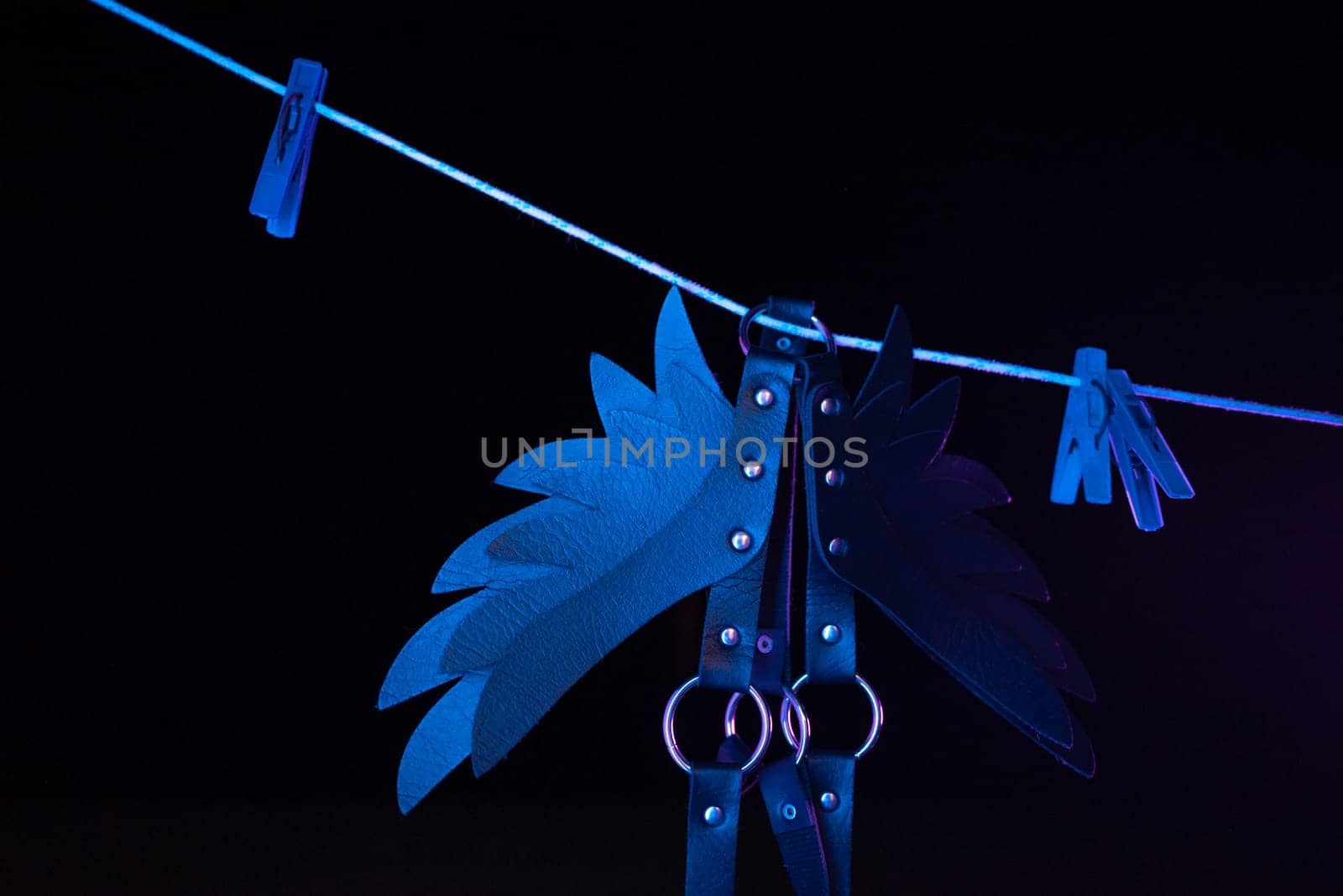 bdsm leather shoulder straps in the shape of wings hanging on clothesline in neon light on a black background, conceptual photos