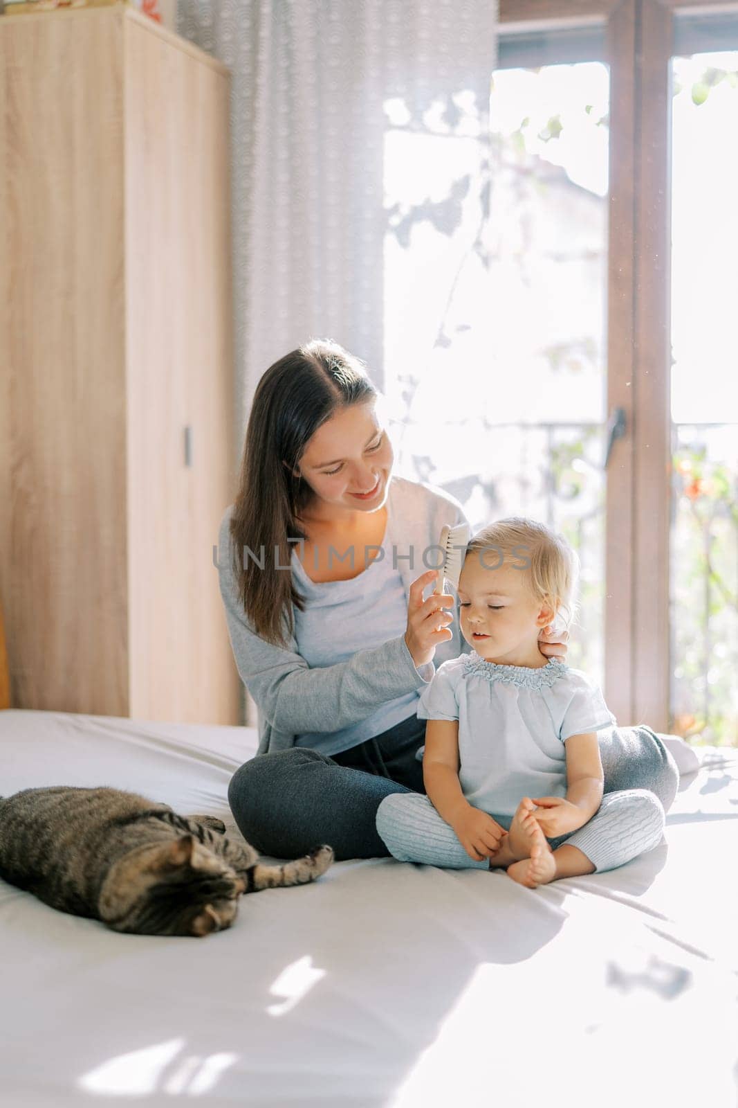 Smiling mother combing little girl hair with a wide brush while sitting on the bed with her. High quality photo