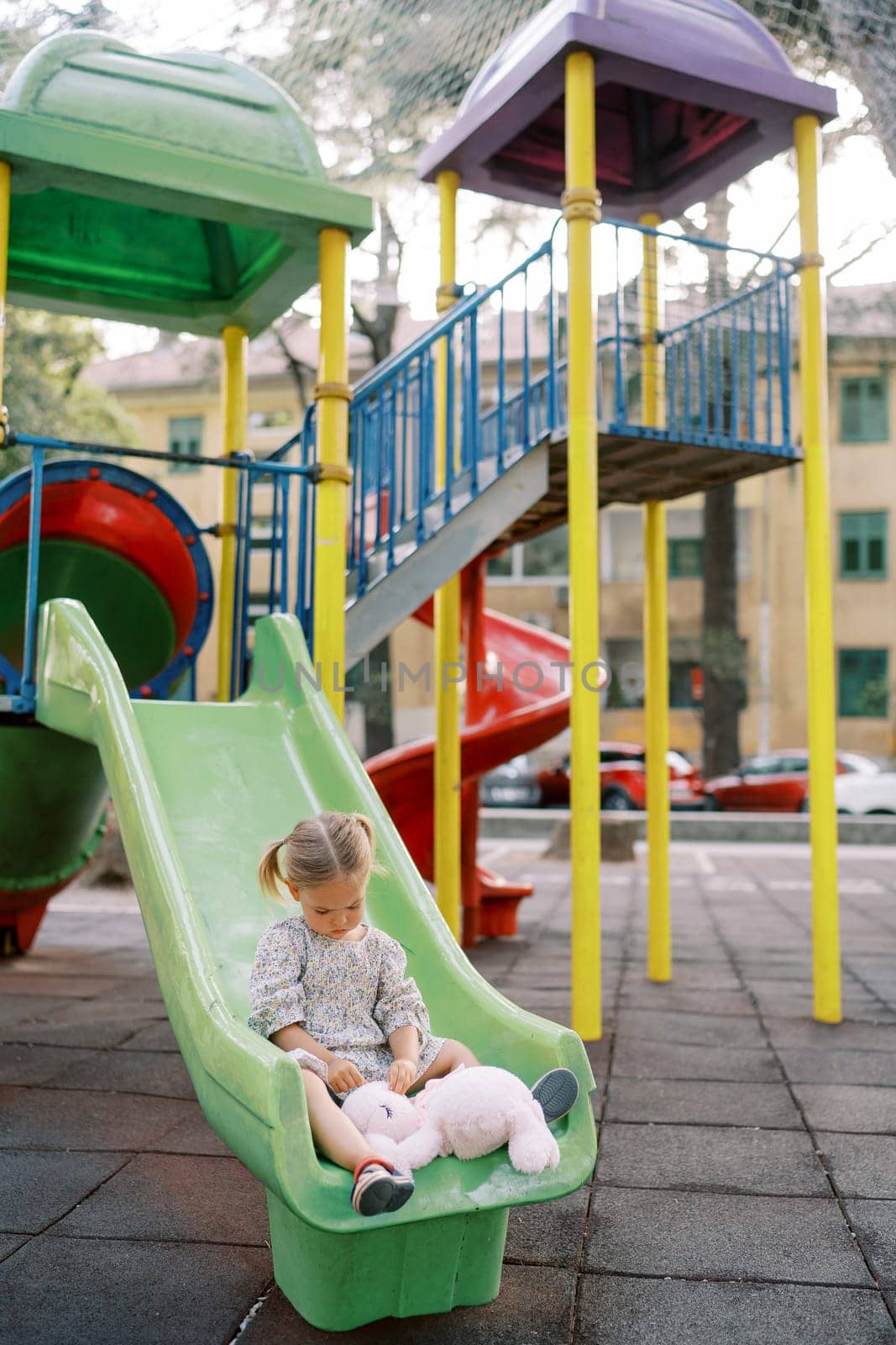 Little girl sits on a colorful slide at the playground and plays with a plush bunny. High quality photo