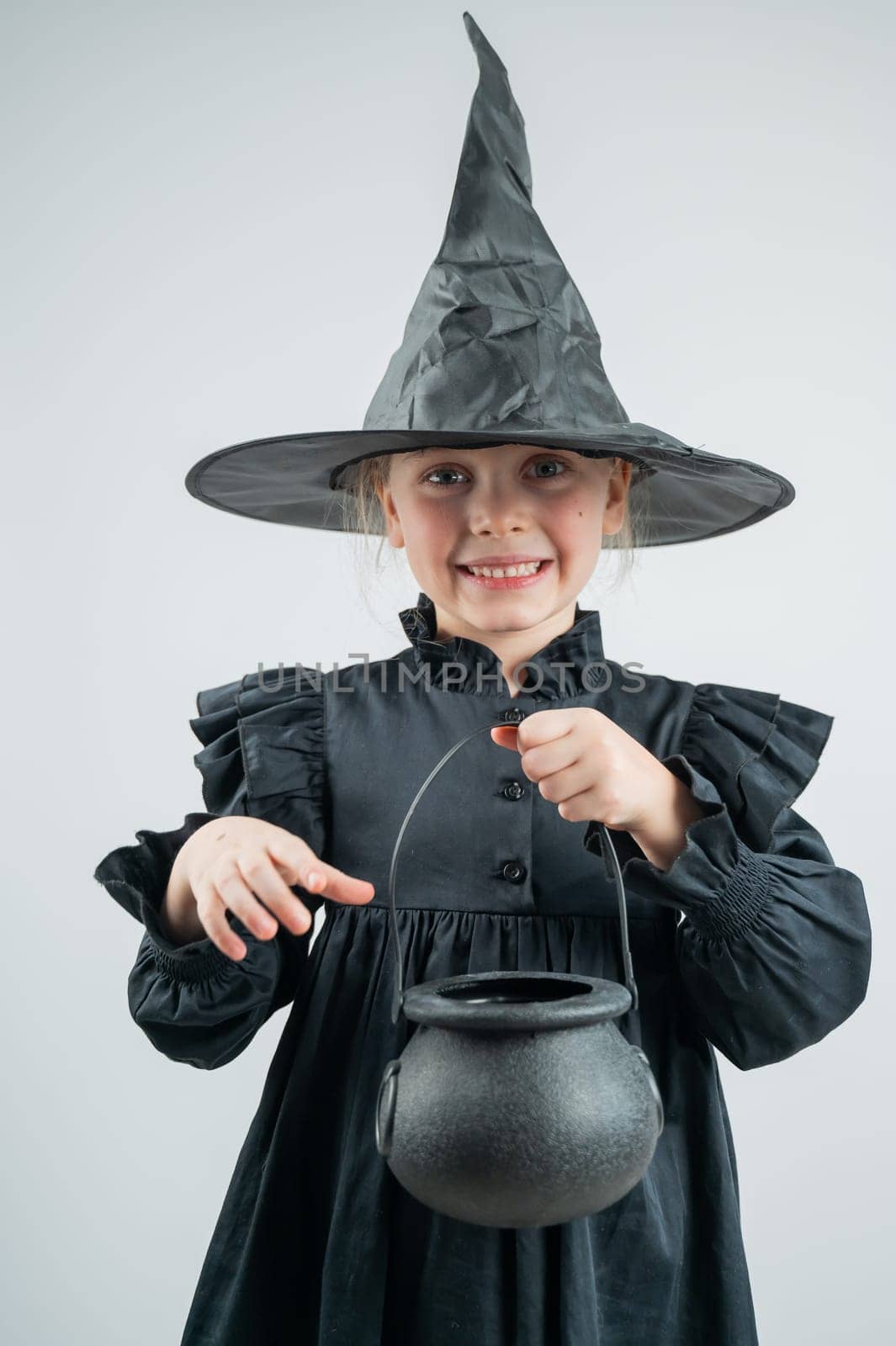 Portrait of a little Caucasian girl in a witch costume holding a cauldron on a white background. Vertical photo