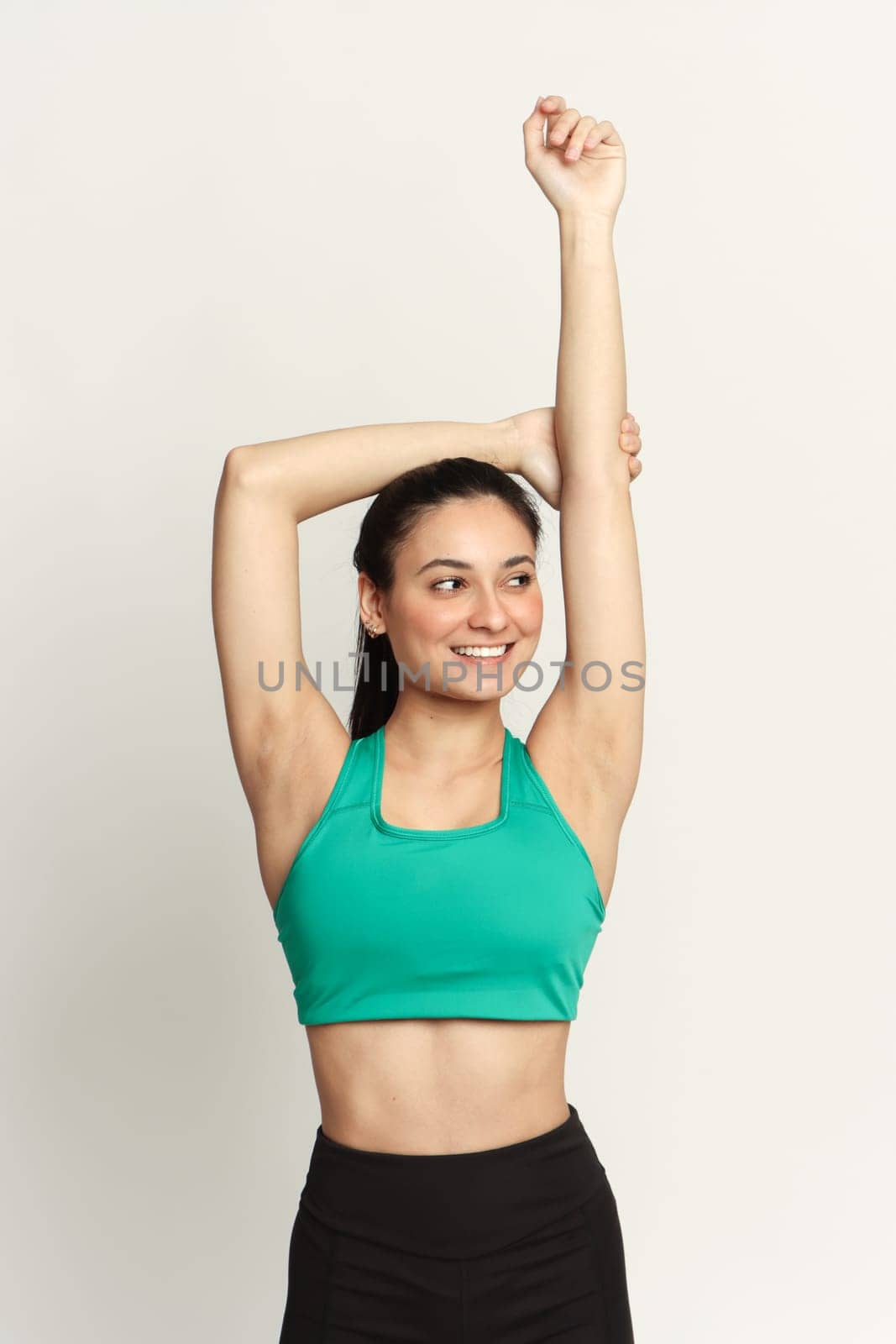 A young woman dresses in sports clothes, raising arms over her head, wears a green top tank, smiling, against a white backdrop in studio.