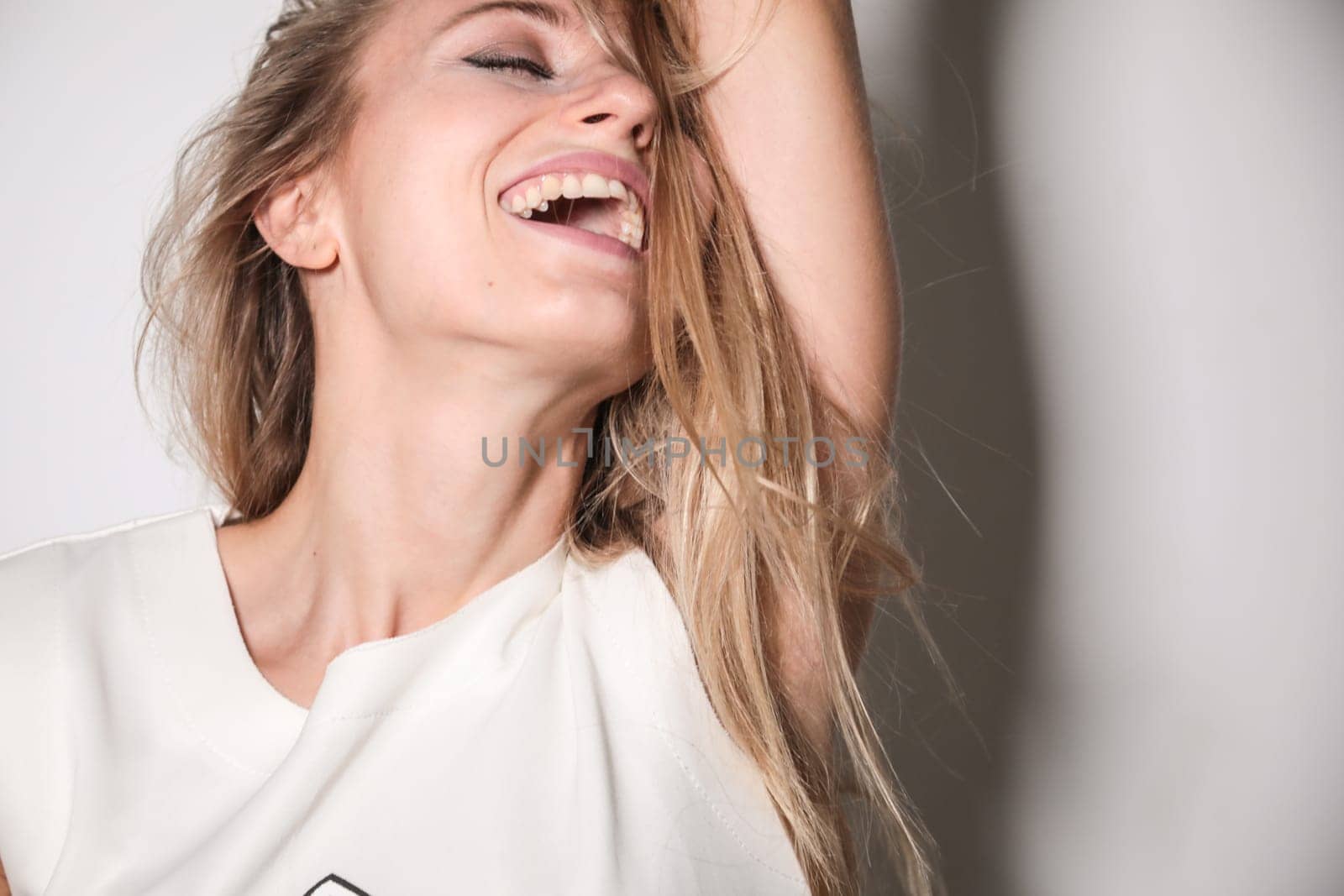 A young model, blonde, dressed in a T-shirt with trimmed sleeves, placed in front of a white wall and illuminated by a strong flash light, laughs out laughing with her eyes closed, resting her head on her arm.
