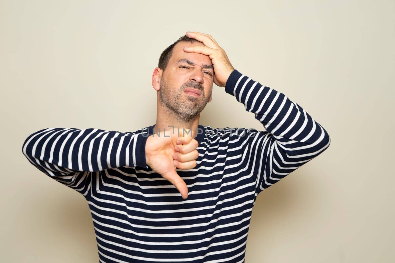 Hispanic man with beard about 40 years old wearing striped sweater looking unhappy and angry showing rejection and negative with thumbs down gesture. isolated on beige studio background. by Barriolo82