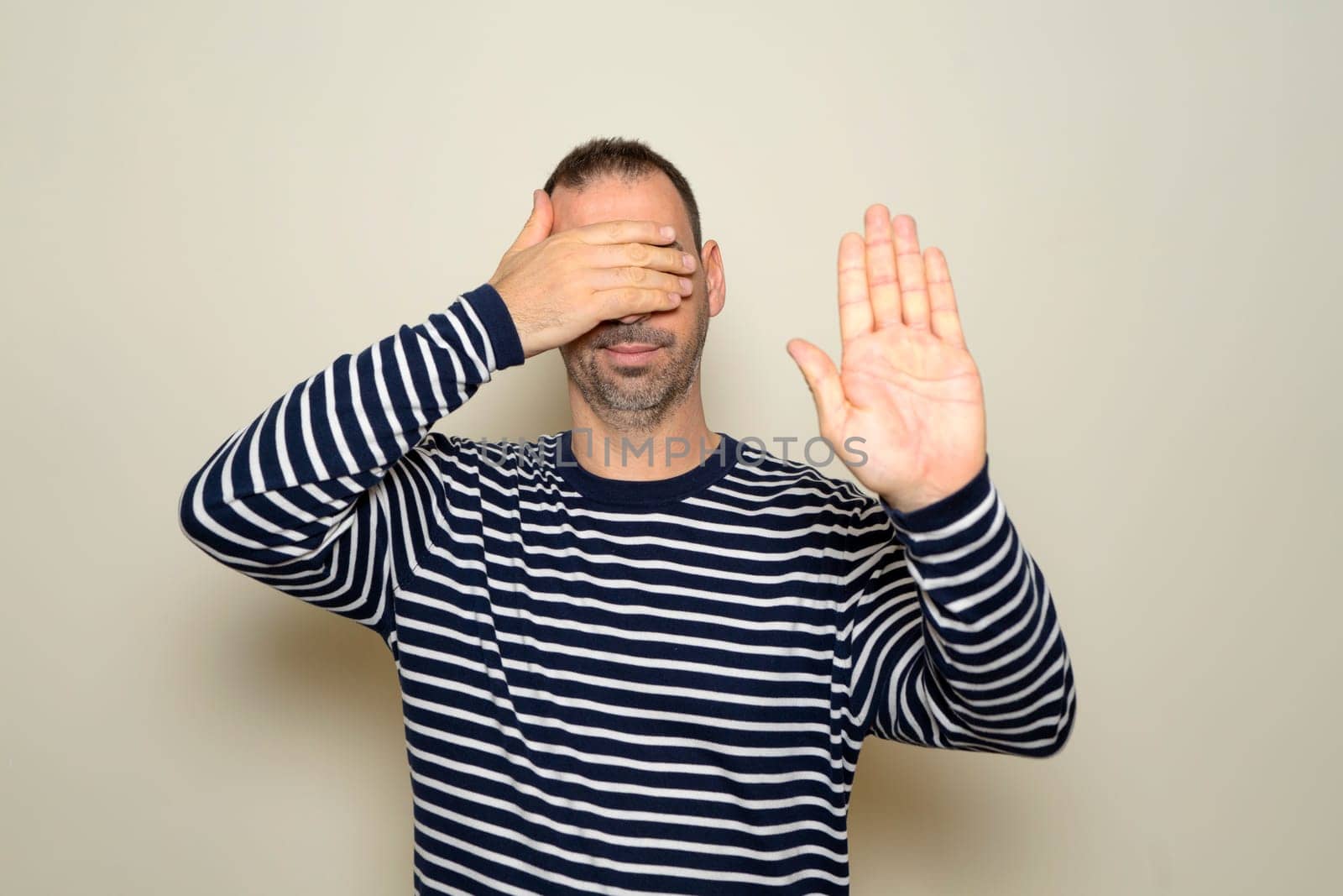 Hispanic man with beard in his 40s wearing a striped sweater covering his eyes with his hands and making stop gesture with a sad and afraid expression. Embarrassed and negative concept.