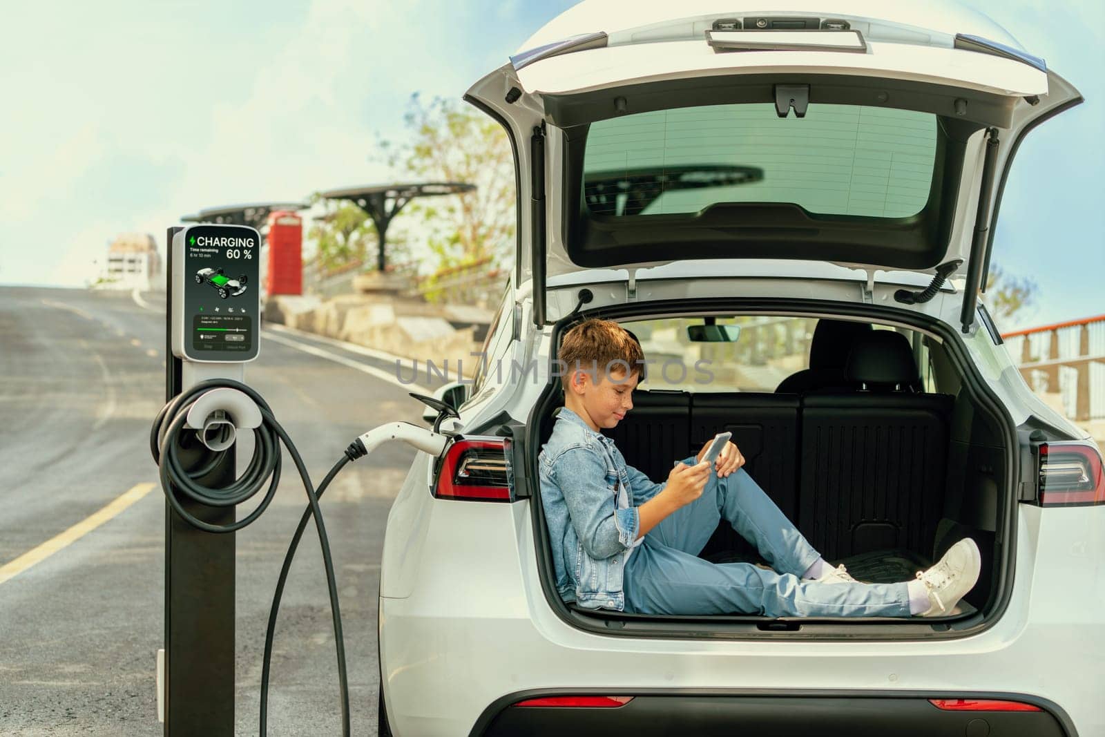 Little boy sitting on car trunk, using smartphone while recharging eco-friendly car from EV charging station. EV car road trip travel as alternative vehicle using sustainable energy concept. Perpetual