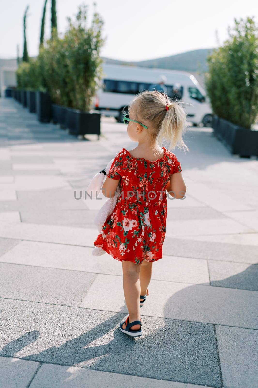 Little girl walks with a pink toy through a square with green trees in flowerpots and looks to the side. Back view. High quality photo
