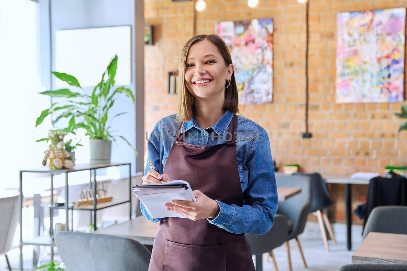 Successful young woman service worker owner in apron with working notepad pen looking at camera in restaurant cafeteria coffee pastry shop interior. Small business staff occupation entrepreneur work