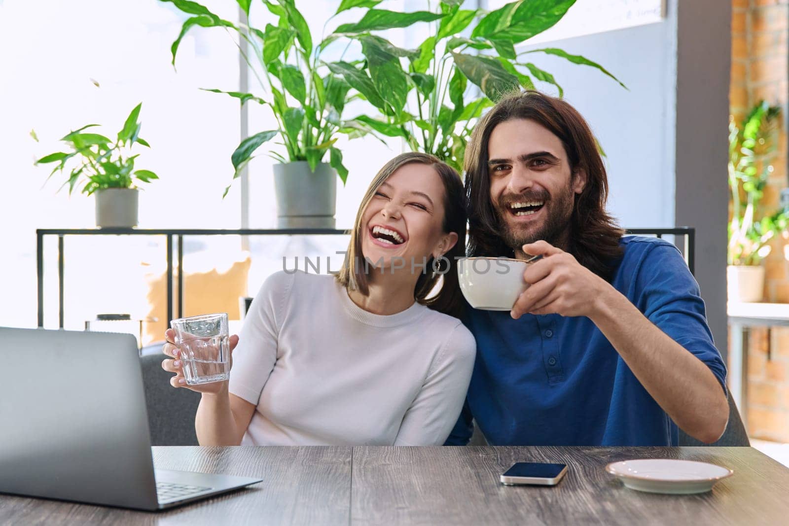 Happy joyful laughing young couple looking at laptop together while sitting in cafeteria. Leisure time for two, communication, togetherness, relationship, lifestyle, work, study remotely