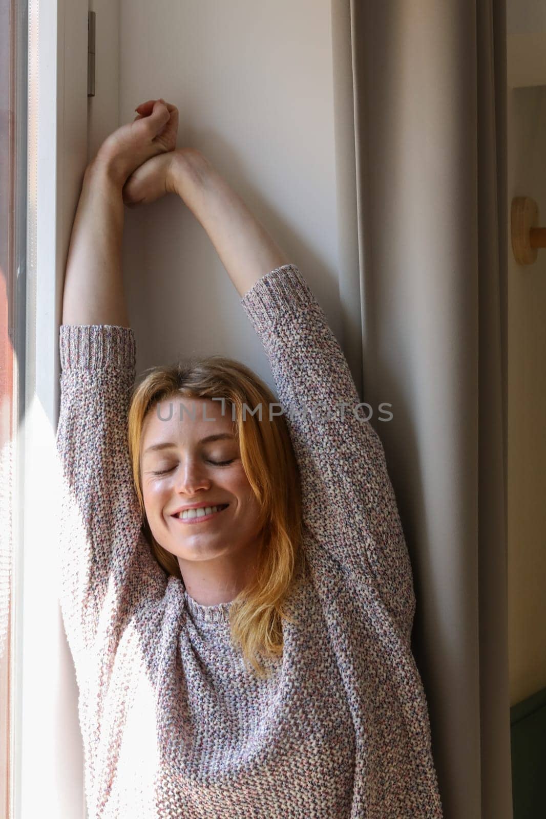 A young woman is streteching her arms, opver her head, resting on the window, with the sun on her right, in her room.