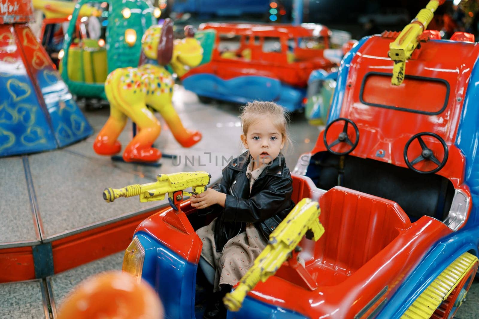 Little girl sits in a toy car on a carousel, holding the steering wheel-gun. High quality photo