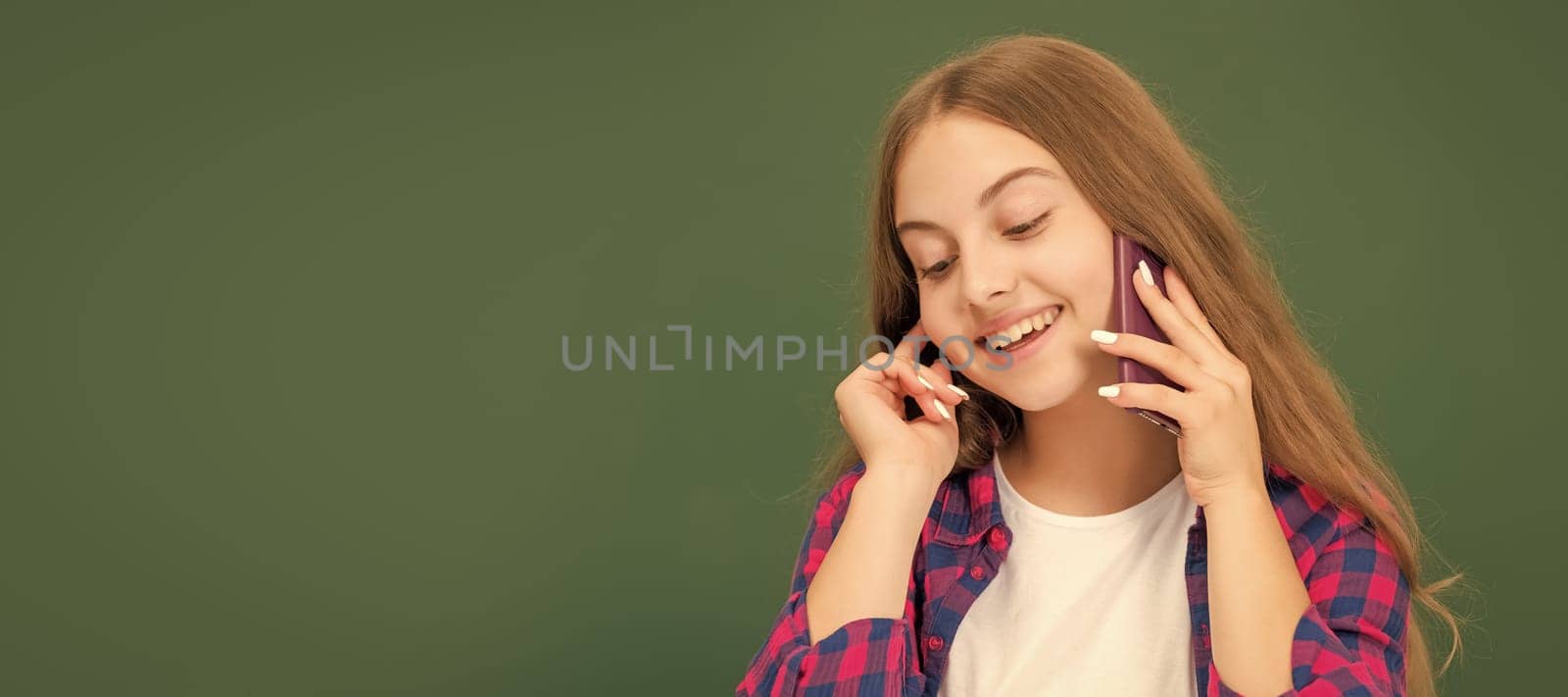 happy kid speaking on smartphone in classroom at blackboard, copy space, conversation. Horizontal isolated poster of school girl student. Banner header portrait of schoolgirl copy space