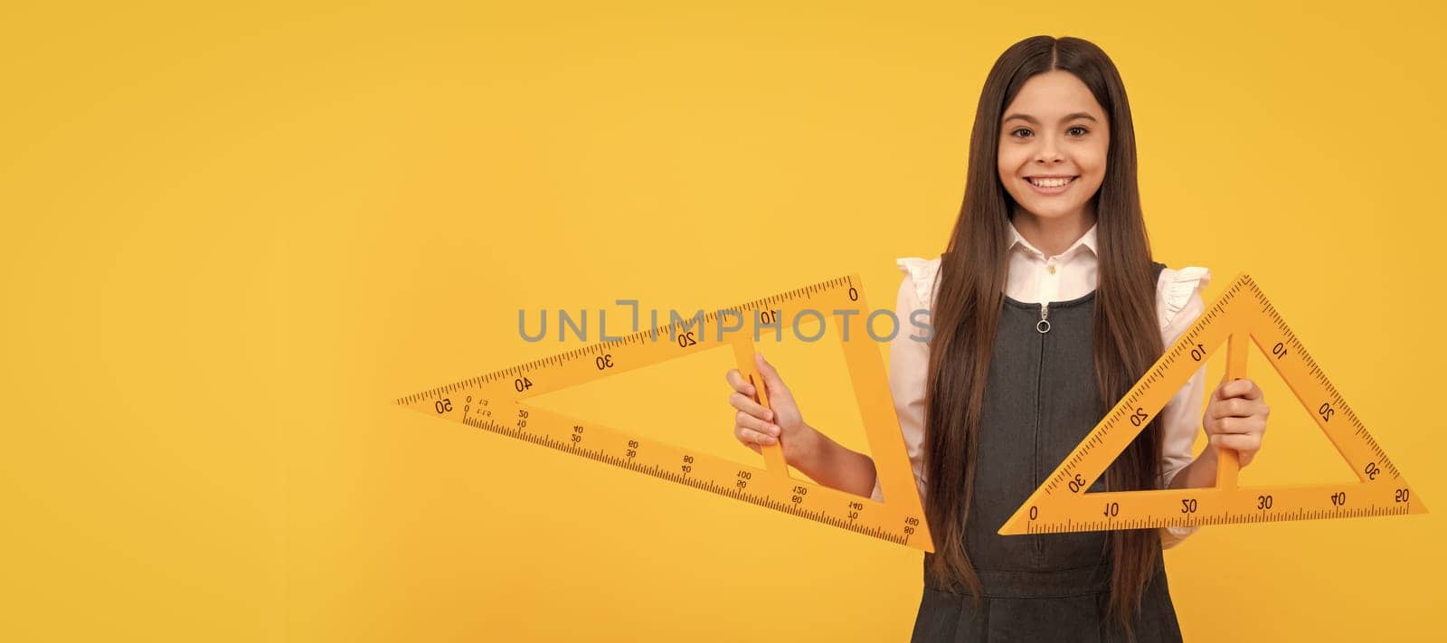 cheerful teen girl hold math triangle ruler in school on yellow background, education. Banner of school girl student. Schoolgirl pupil portrait with copy space. by RedFoxStudio