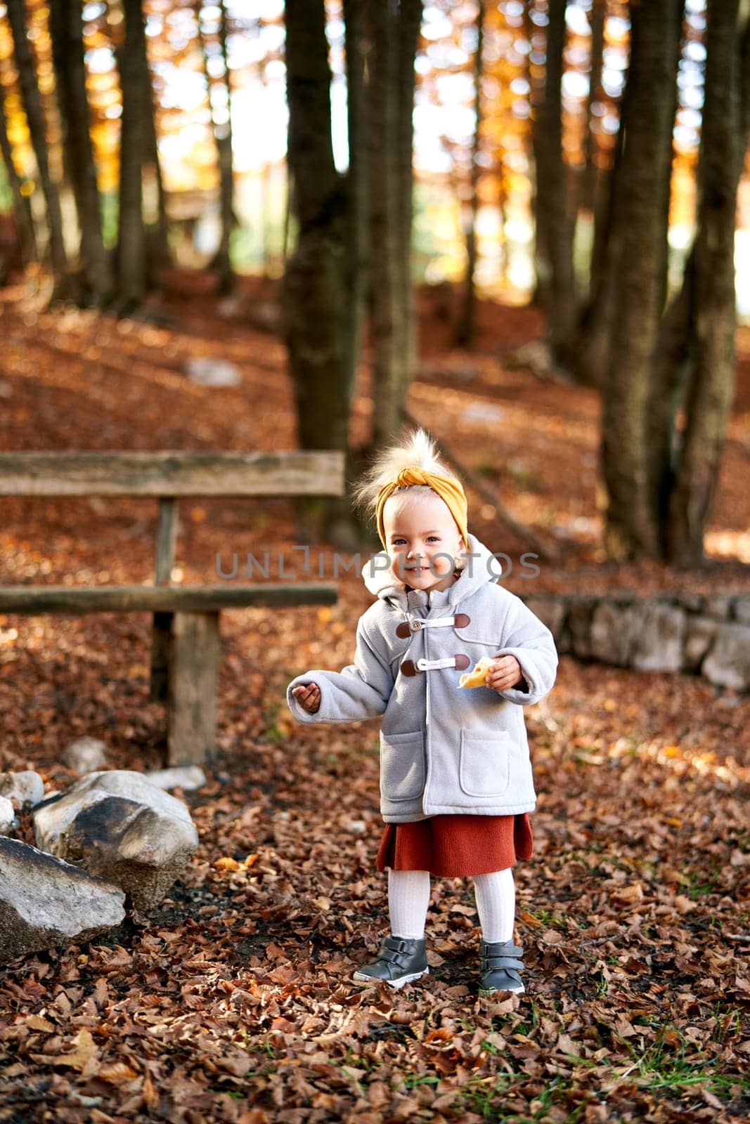Little girl with a bun in her hand stands on fallen leaves near a park bench. High quality photo