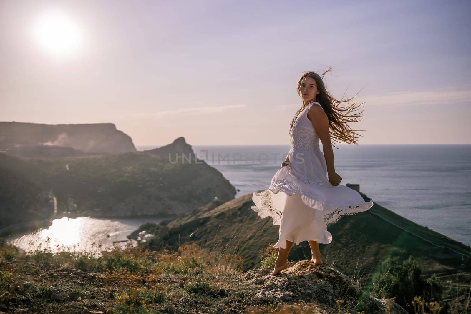 A woman stands on a hill overlooking the ocean. She is wearing a white dress and has long hair. The scene is serene and peaceful, with the sun shining brightly in the background. by Matiunina