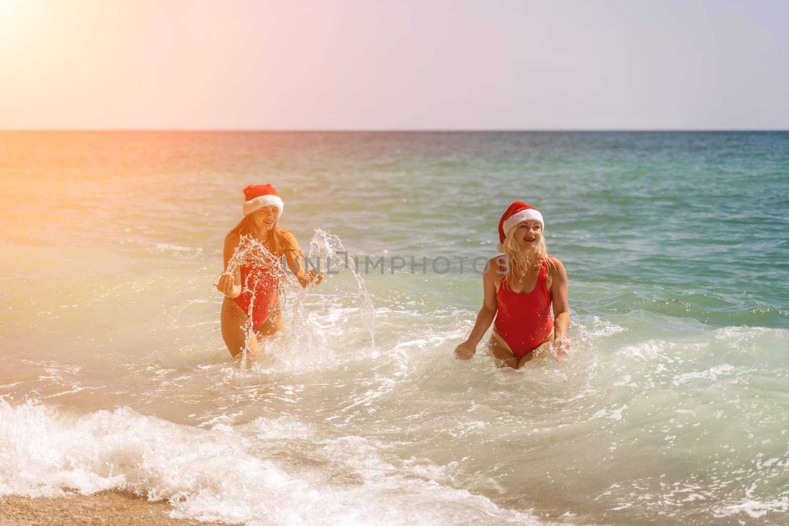 Women Santa hats ocean play. Seaside, beach daytime, enjoying beach fun. Two women in red swimsuits and Santa hats are enjoying themselves in the ocean waves. by Matiunina
