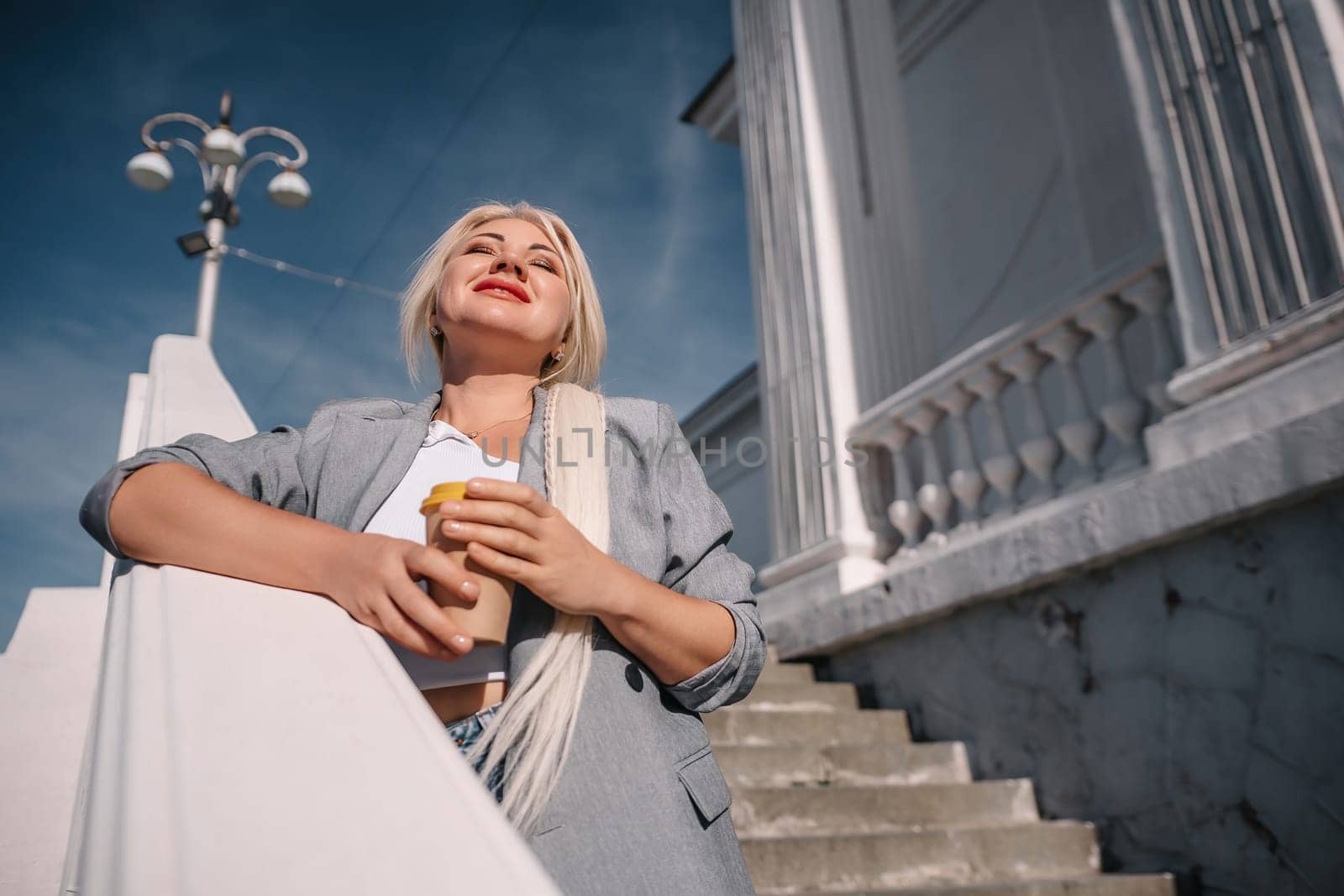A blonde woman in a suit is holding a coffee cup and looking up at the sky. Concept of sophistication and elegance, as the woman is dressed in a business suit and holding a coffee cup