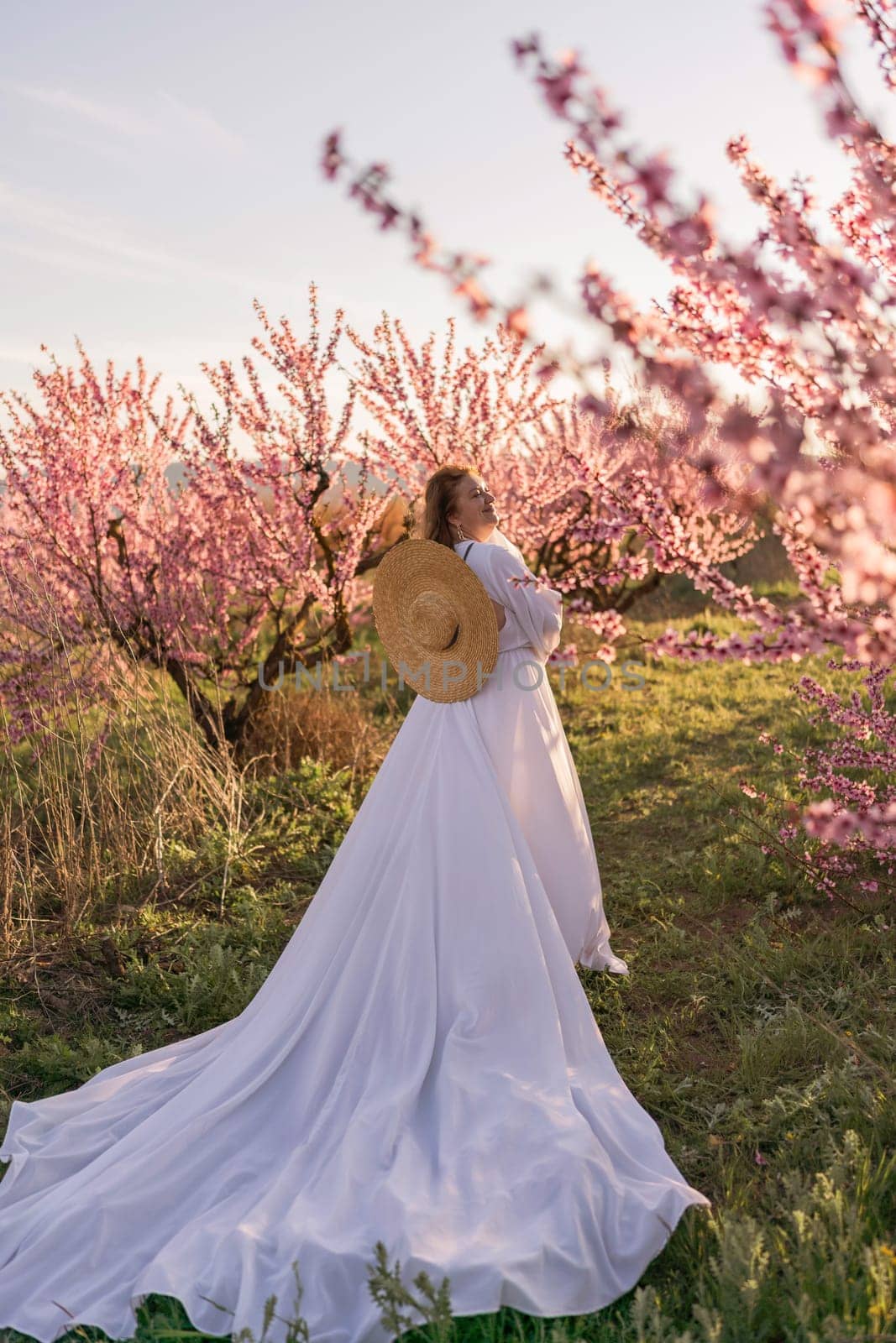 Woman blooming peach orchard. Against the backdrop of a picturesque peach orchard, a woman in a long white dress and hat enjoys a peaceful walk in the park, surrounded by the beauty of nature. by Matiunina