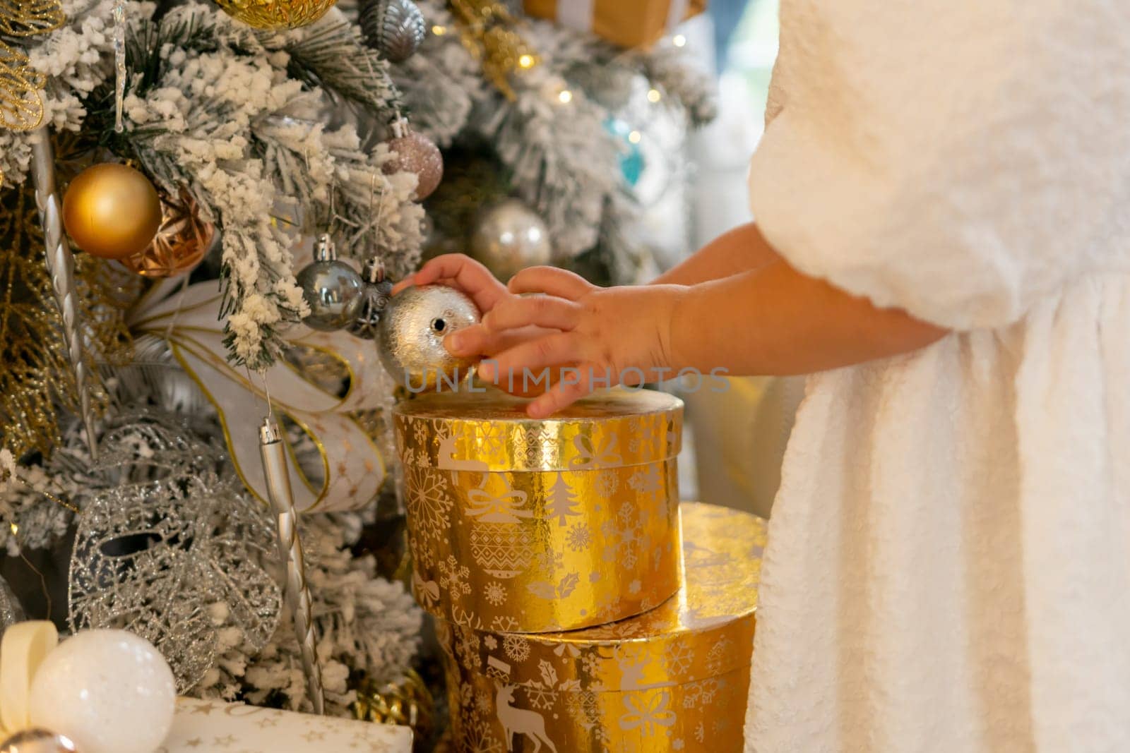 A woman is opening a gold box with a silver ball inside. The box is decorated with gold and silver ornaments, and the woman is wearing a white dress. The scene is set in a room with a Christmas tree
