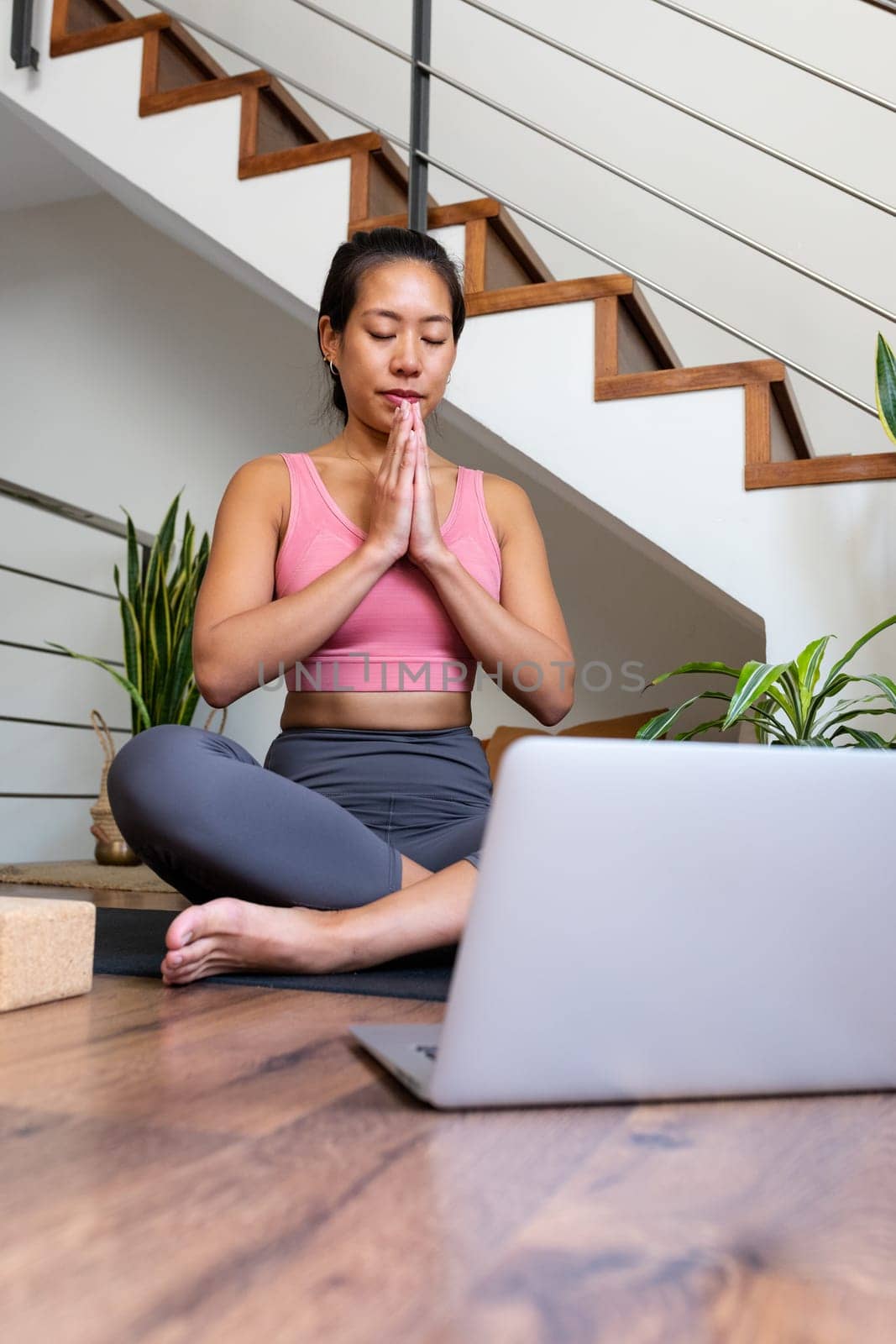 Vertical portrait of young Asian woman doing online meditation with hands in prayer at home living room sitting on yoga mat using laptop. Spirituality concept.