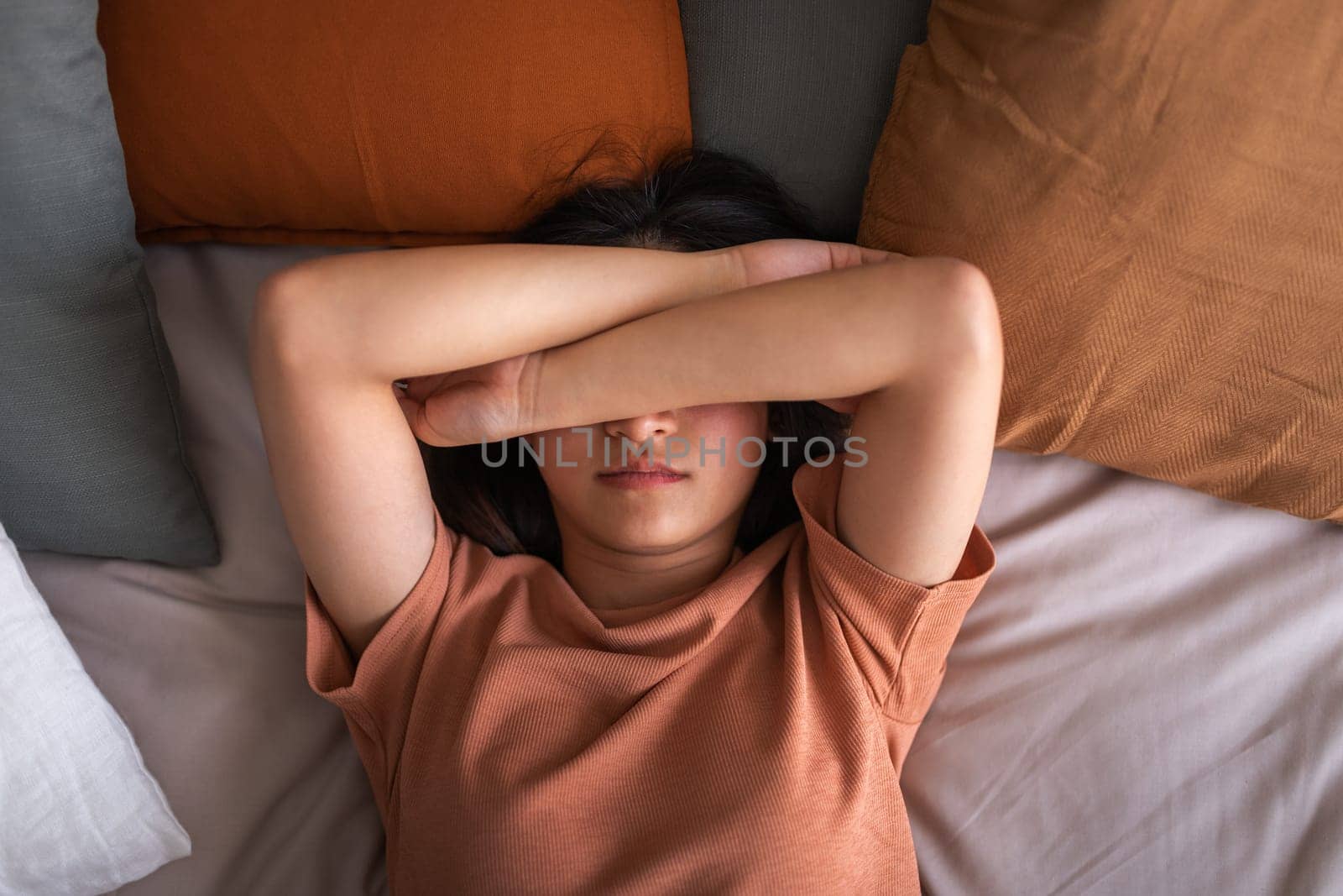 Top view of depressed, sad young asian woman lying on bed covering eyes with arms. Depression, mental health concepts.