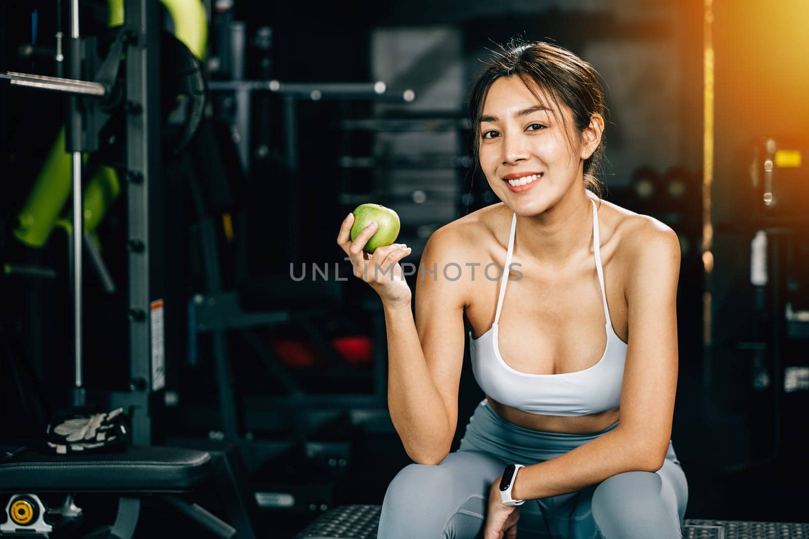 A portrait of a young Asian woman smiling as she holds a green apple in a fitness gym, emphasizing the importance of a balanced diet for achieving health and fitness. Clean food and lifestyle Healthy