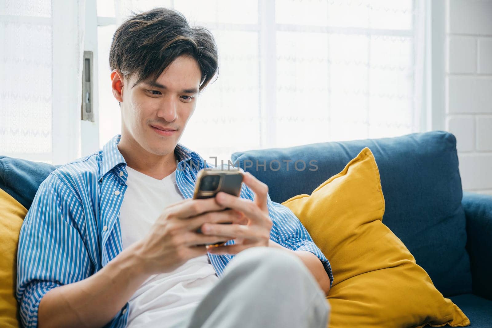 A young man smiling and talking on his phone while sitting comfortably on a sofa. Engaged in successful communication and technology he exudes happiness and relaxation. scrolling on social media.