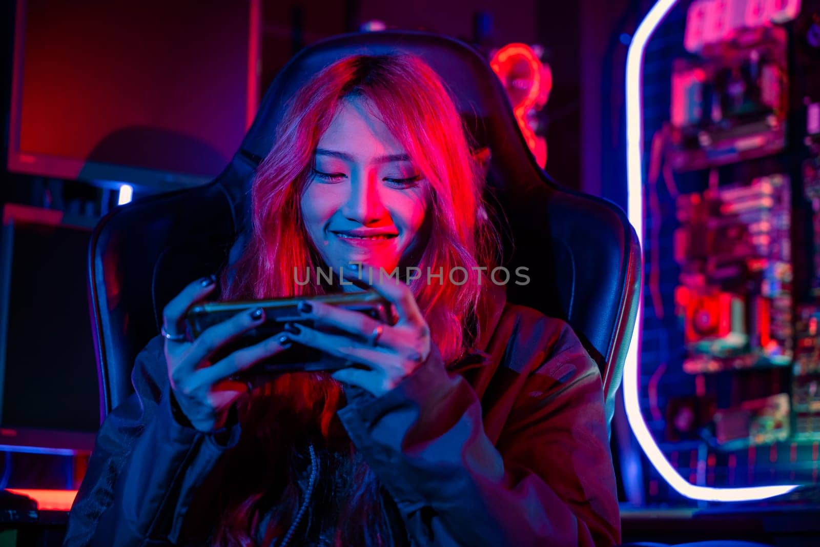Gamer playing online game application on mobile phone wear gaming headphones, Asian woman live stream she play video game via smartphone at home neon lights living room