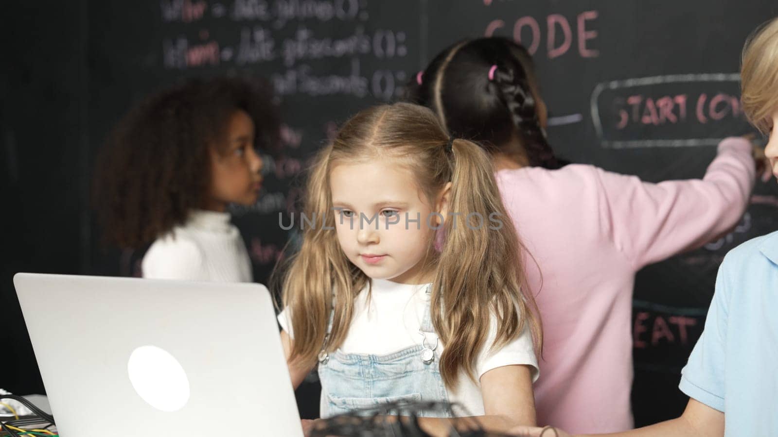 Smart girl using laptop programing engineering code with diverse friends ay table with microchip and main board scatter around in STEM classroom at blackboard written with coding prompt. Erudition.