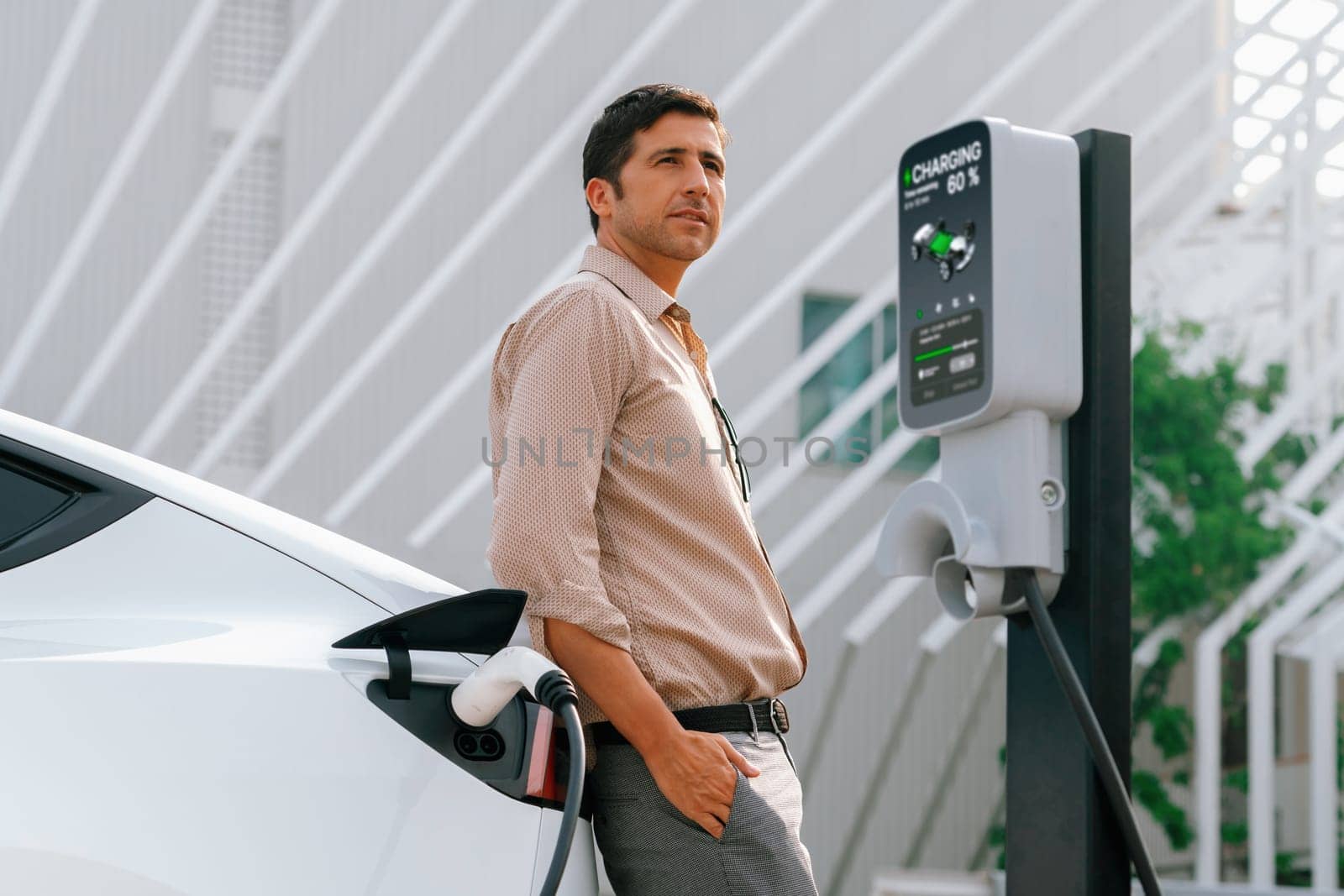 Young man put EV charger to recharge electric car's battery from charging station in city commercial parking lot. Rechargeable EV car for sustainable environmental friendly urban travel. Expedient