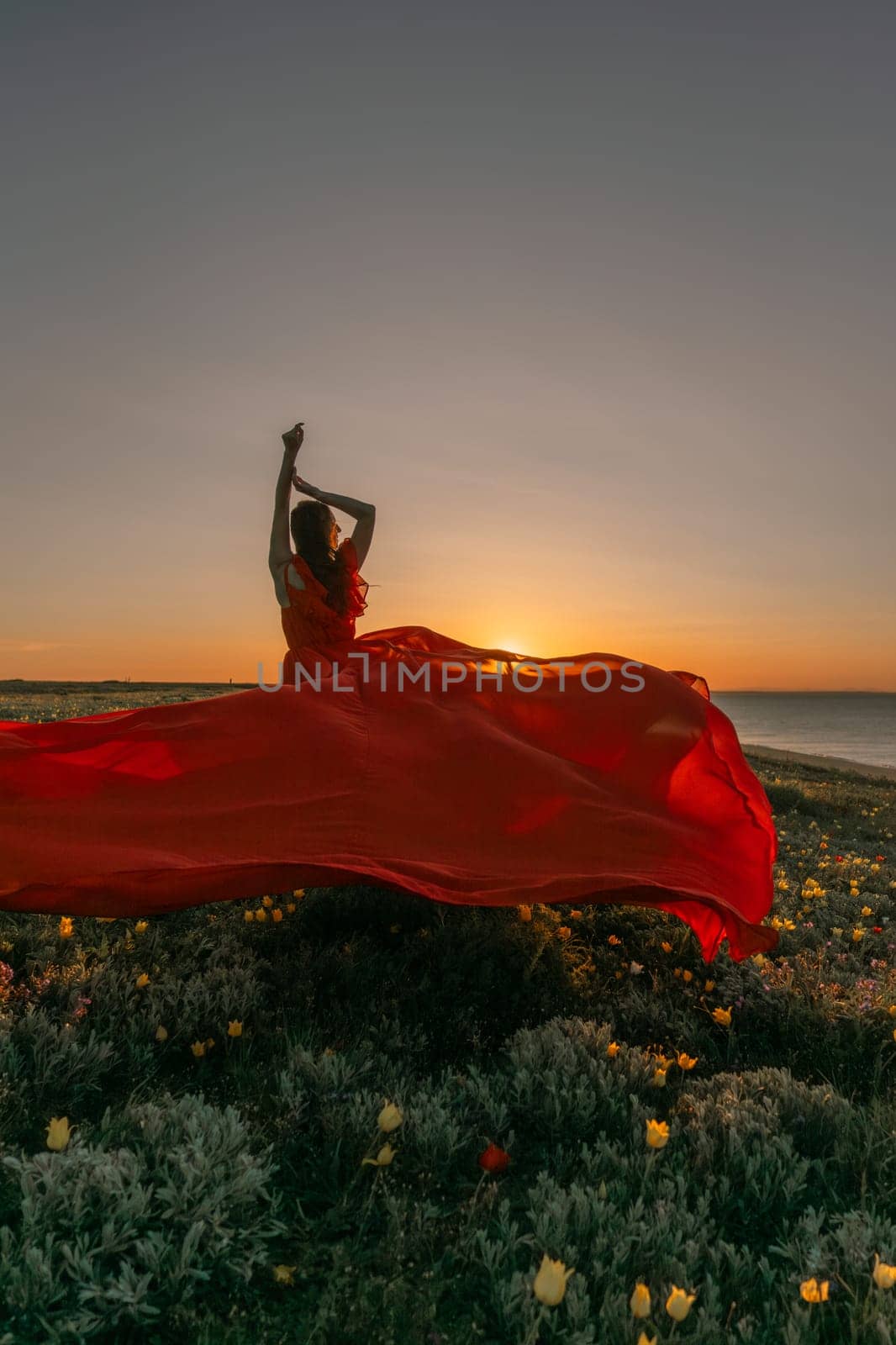 A woman in a red dress is standing in a field with the sun setting behind her. She is reaching up with her arms outstretched, as if she is trying to catch the sun. The scene is serene and peaceful. by Matiunina