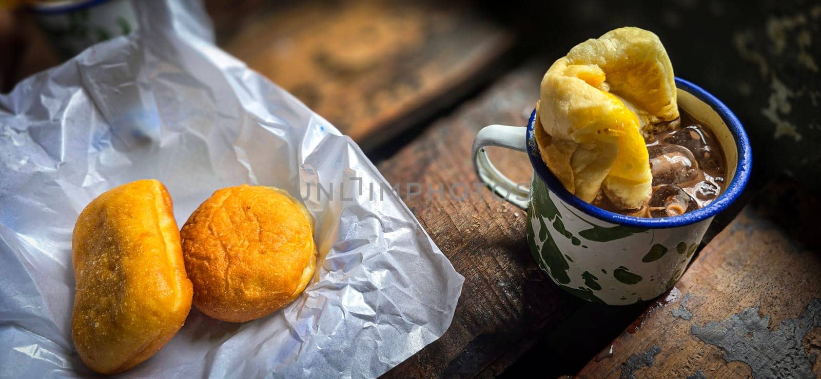 Ice chocolate drink on a vintage Indonesian iron mug with random green pattern called Blirik cup or cangkir Blirik on bokeh background along with chocolate filling bread on a cafe in Bogor